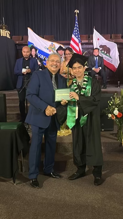 Graduation ceremony with a student in a black cap and gown with an Aloha Lei, holding a diploma, shaking hands with the principal.