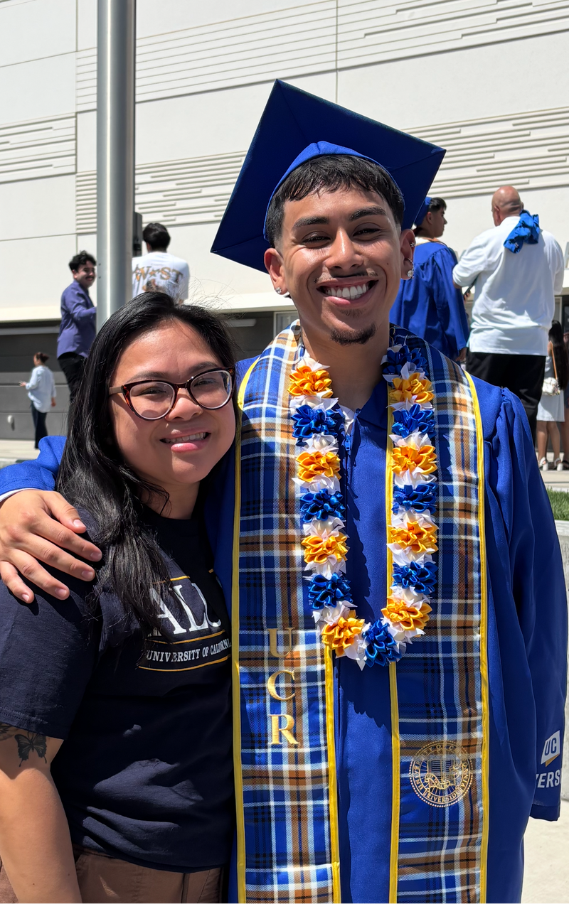Male graduate in blue cap and gown wearing a blue and yellow aloha lei, standing with his arm around his friend.
